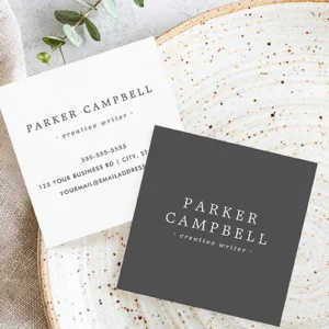 Front and back of a customizable square business card. The front has a dark charcoal background with centered name and title in all-caps serif and smaller sans-serif italics fonts. The back is plain white, repeating the centered name and description fields in dark text, with custom contact info such as phone, address, and website/email details stacked below.