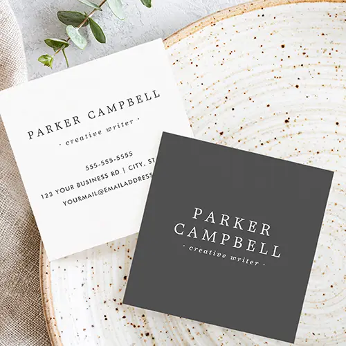 Front and back of a customizable square business card. The front has a dark charcoal background with centered name and title in all-caps serif and smaller sans-serif italics fonts. The back is plain white, repeating the centered name and description fields in dark text, with custom contact info such as phone, address, and website/email details stacked below.