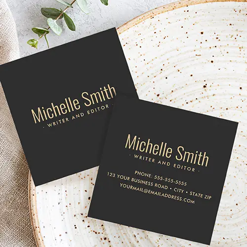 Square business card with a solid, very dark gray background. Centered on the card is the name printed in a warm yellow tone sans-serif font. Directly below the name is a smaller line of custom text in the same color, suggesting a job title or tagline.
