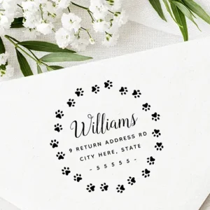 Round rubber stamp design showing a circular wreath border made of small paw prints surrounding a central flowing script name and three lines of uppercase address typography flanked by dashes.