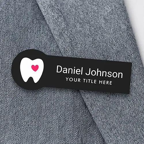 Rectangular black plastic name badge featuring a rounded circular left tab with a white tooth and pink heart icon, alongside custom white employee name and title text lines.