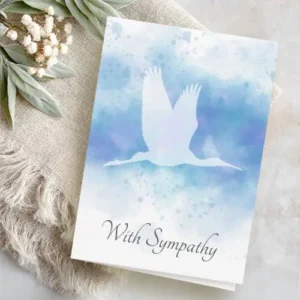 A folded white card, featuring a central gradient watercolor wash of blue and lavender. At the center is a light silhouette of a crane in mid-flight. Delicate water spatters are visible. Customizable text "With sympathy" on the front and message inside.