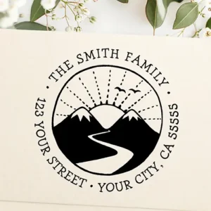 Circular self-inking stamp graphic featuring twin snow-capped mountains, a winding river, dashed sunbeams, and curved custom family address typography.