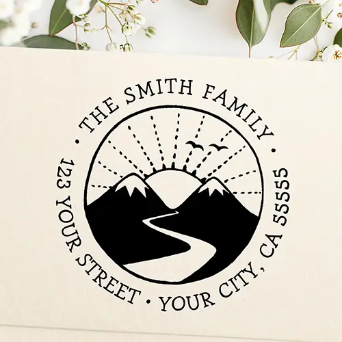 Circular self-inking stamp graphic featuring twin snow-capped mountains, a winding river, dashed sunbeams, and curved custom family address typography.