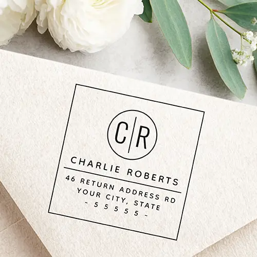 Custom rubber stamp featuring a circular split monogram emblem at the top, followed by a bold custom name, a horizontal divider line, and clean address text lines at the bottom.