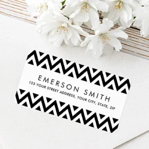 Horizontal rectangular white address sticker featuring bold black and white zigzag chevron pattern borders along top and bottom with centered black sans-serif text for name and address.