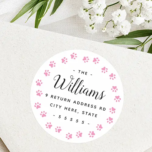 Round white adhesive label surrounded by a border of pink paw prints, featuring black calligraphic script and custom address typography in the center.