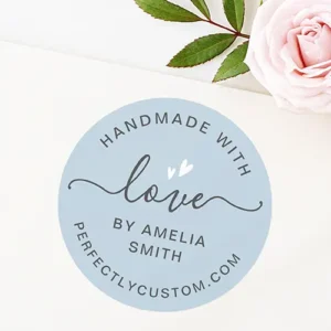 A circular sticker featuring a soft dusty blue background, a charcoal script word "love" with white hearts, and curved template text for a maker name and website.
