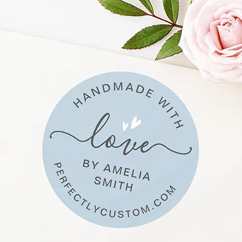 A circular sticker featuring a soft dusty blue background, a charcoal script word "love" with white hearts, and curved template text for a maker name and website.