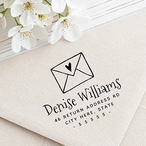 Rubber stamp design featuring an illustrated envelope with a heart motif, a playful handwritten script name, two lines of uppercase address text, and a dashed ZIP code row.