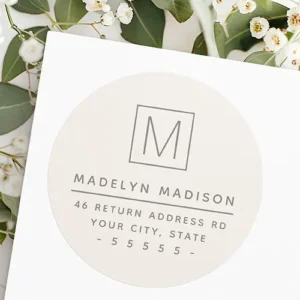 A circular sticker label on a pale taupe brown background featuring taupe grey typography. At the top is a square outline enclosing a single letter monogram, followed by a full name line, a thin horizontal divider line, and three address text lines below.