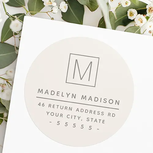 A circular sticker label on a pale taupe brown background featuring taupe grey typography. At the top is a square outline enclosing a single letter monogram, followed by a full name line, a thin horizontal divider line, and three address text lines below.
