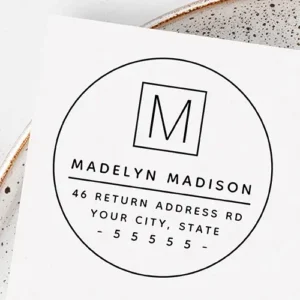 Circular rubber stamp design showing a single letter inside a square frame at the top, followed by a full custom name, horizontal line divider, and structured address text.