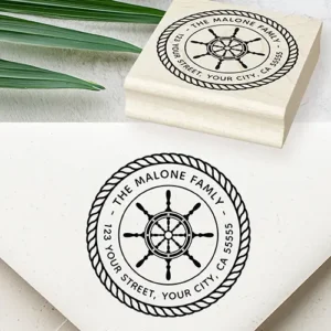 Round rubber stamp design featuring a central ship's wheel surrounded by arched uppercase custom name and address text, enclosed within a twisted nautical rope border.