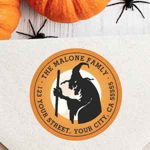 Circular pumpkin-orange return address sticker displaying a central black silhouette of a wicked witch holding a staff in front of a full moon, bordered by arched black typewriter text.