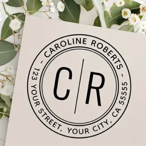 Round rubber stamp design showing a central split initial monogram divided by a vertical line, enclosed by double circular ring borders and curved custom name and address text.