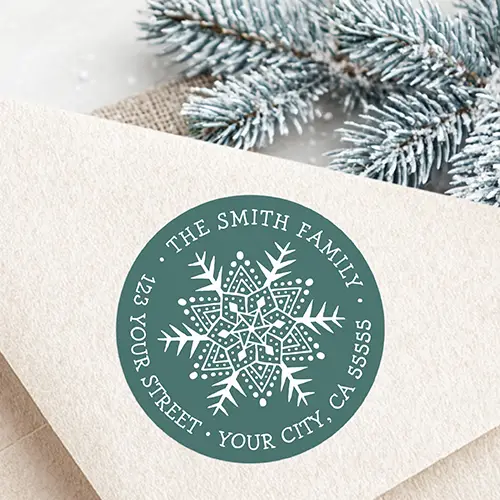 Round pine green return address sticker featuring a whimsical folk art style white snowflake illustration at the center, surrounded by curved uppercase serif text displaying a family name and complete address.