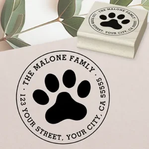 Round rubber stamp design showing a large central solid paw print graphic surrounded by curved typewriter font displaying a custom family name and address details within a circular frame.