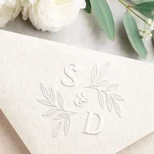 A 3D embossed paper envelope displaying two letter initials arranged diagonally with a centered ampersand, framed by two decorative branching leaf sprigs with detailed leaf outlines. Monogram embosser plate for wedding invitations & couples correspondence.