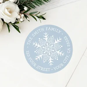 Round pastel dusty blue return address sticker featuring a detailed white snowflake illustration at the center surrounded by curved uppercase serif text displaying a family name and complete address.