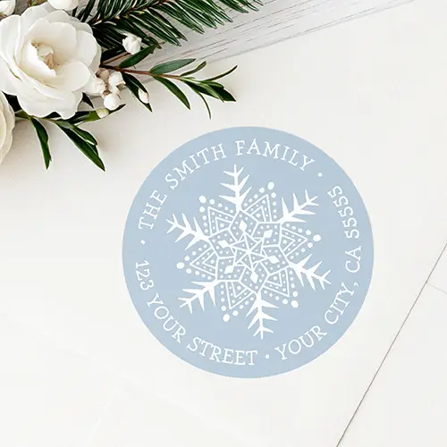 Round pastel dusty blue return address sticker featuring a detailed white snowflake illustration at the center surrounded by curved uppercase serif text displaying a family name and complete address.