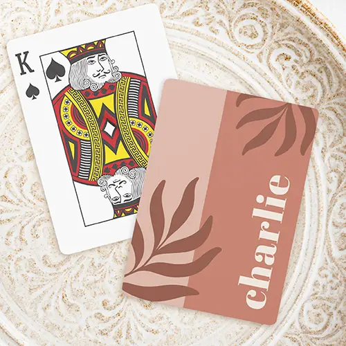 A rectangular custom playing card with rounded corners, featuring a vertically split background of dusty blush pink brown on the left and earthy terracotta on the right. Dark brown abstract leaf fronds decorate the opposite corners, and a custom name is written vertically on the right side in a thick, cream-colored serif font.