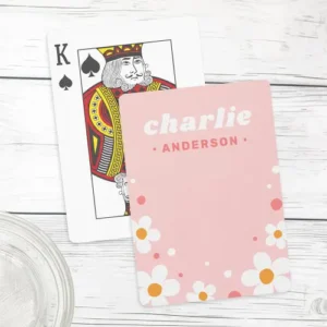 Custom playing cards in pink, featuring a personalized name at the top and a border of white daisies with orange centers at the bottom. Decorative white and pink circles are scattered around the flowers.