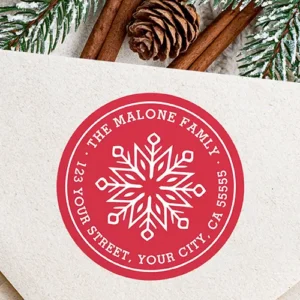 Round red holiday return address sticker featuring a central white geometric snowflake motif encircled by arched white custom name and address text within a thin white border ring.