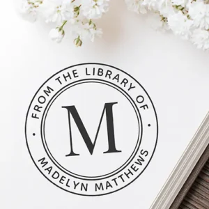 Round self-inking stamp featuring a large central serif letter M enclosed by a double circular border and curved custom library owner text.
