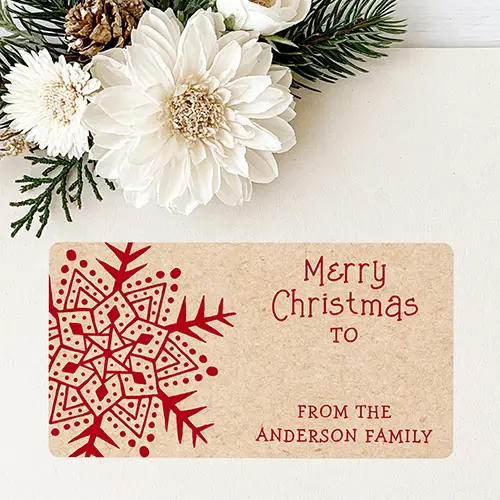 Rectangular sticker label with kraft paper background featuring a detailed dark red snowflake on the left, Merry Christmas typography with a TO line, and a custom family signature at the bottom right.