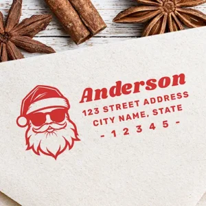 Rubber stamp design featuring a graphic Santa Claus illustration with sunglasses on the left alongside a bold italic custom surname and three lines of uppercase address typography flanked by hyphens on the right.