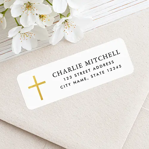 Horizontal rectangular white address sticker featuring a printed faux gold cross on the left and black serif name and sans-serif address text on the right.