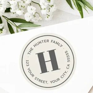 Circular off-white return address label sticker featuring a large grey serif monogram letter in the center framed by a circular grey border and curved dark grey family name and address text.