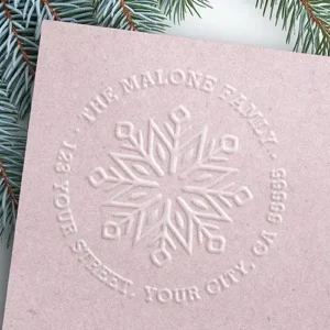 A raised 3D paper embosser seal on textured paper. The center displays an intricate geometric snowflake, surrounded by a circular border of arched serif text with a custom family name along the top and street address along the bottom.