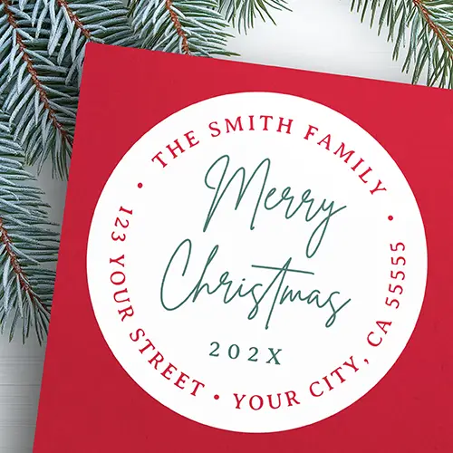A circular self-adhesive sticker with a white background featuring a handwritten green script reading Merry Christmas in the center with a custom year line below. A circular outer ring of red uppercase serif text displays a family name on top and a return address on the bottom.