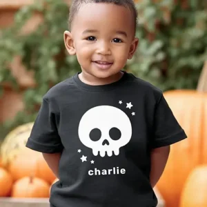 A black short-sleeve toddler t-shirt displaying a white graphic illustration of a friendly cartoon skull with round eye sockets, surrounded by small white stars and a personalized name placeholder below in bold white lowercase text.