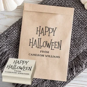 A square Halloween rubber stamp. The top section reads "HAPPY HALLOWEEN" in a tall, stylized gothic serif font, followed by "FROM" and a customizable full name in spaced all-caps serif typeface below.