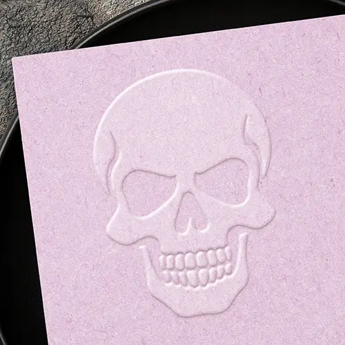A 3D embossed skull seal on textured paper showcasing an outlined human skull shape with detailed eye sockets, nasal cavity, cheek structure, and individual teeth.