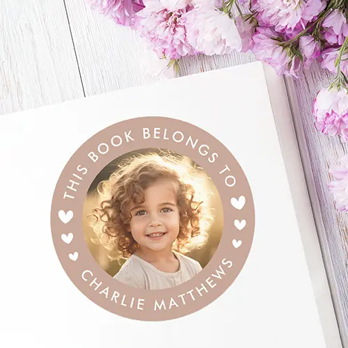 A personalized round bookplate sticker featuring a central custom photo area), a light brown border, white arched text 'THIS BOOK BELONGS TO' and your custom name. Decorative white hearts on the sides.