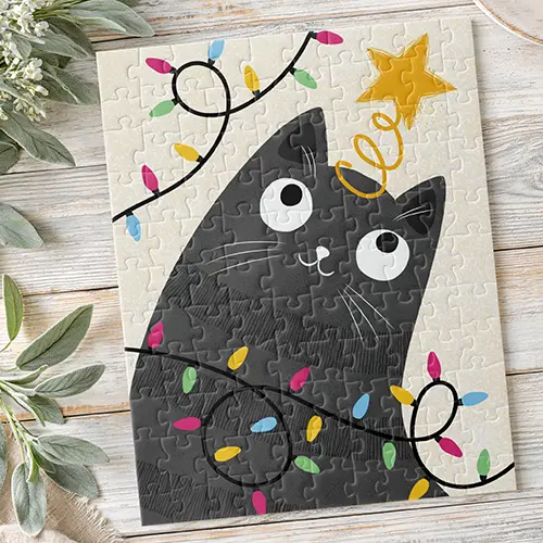 A vibrant jigsaw puzzle featuring a modern illustration of a whimsical, deep gray cat with large white eyes and whiskers. The cat is looking upwards and is playfully tangled in a wire of multi-colored holiday lights (pink, blue, yellow, green bulbs). A gold star tree-topper sits on its head. The background is a soft, warm cream color with a subtle distressed pattern.