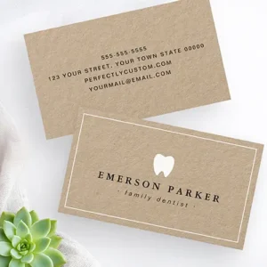 A brown kraft paper business card featuring a small white tooth icon at the top, a name in bold serif font, and custom title or clinic name in italics. A white thin-line border frames the entire design.
