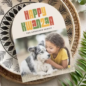 A modern Kwanzaa greeting card with a unique arched shape. The top section is cream-colored and has the words 'HAPPY KWANZAA' in a bold, blocky, multi-colored retro font featuring traditional red, green, and yellow hues. Below the greeting, there is a small custom text line and a rectangular space for a customized family photograph.