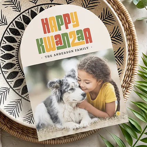 A modern Kwanzaa greeting card with a unique arched shape. The top section is cream-colored and has the words 'HAPPY KWANZAA' in a bold, blocky, multi-colored retro font featuring traditional red, green, and yellow hues. Below the greeting, there is a small custom text line and a rectangular space for a customized family photograph.