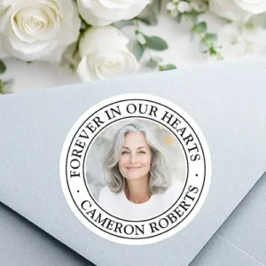 Forever in our hearts white border photo memorial classic round sticker A round white sticker featuring a central circular custom photo field framed by a double black border. The top arc contains black serif text reading "FOREVER IN OUR HEARTS", while the bottom arc displays a custom name, separated by two tiny dot accents.