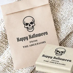 A Halloween rubber stamp design. The top features a detailed skull illustration. Centered below is "Happy Halloween" in a gothic blackletter font, followed by all-caps serif text placeholders reading "FROM" and a customizable name field suitable for family or business details.