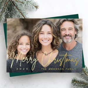 Merry Christmas modern script deep green photo Foil Holiday Card A landscape-oriented custom photo holiday card featuring a full-bleed custom family portrait. Overlaid on the lower half is the phrase "Merry Christmas" in a fluid gold foil script, anchored by gold text fields for the year on the left and a family name on the right.