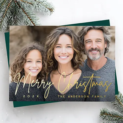 A landscape-oriented custom photo holiday card featuring a full-bleed custom family portrait. Overlaid on the lower half is the phrase "Merry Christmas" in a fluid gold foil script, anchored by gold text fields for the year on the left and a family name on the right.