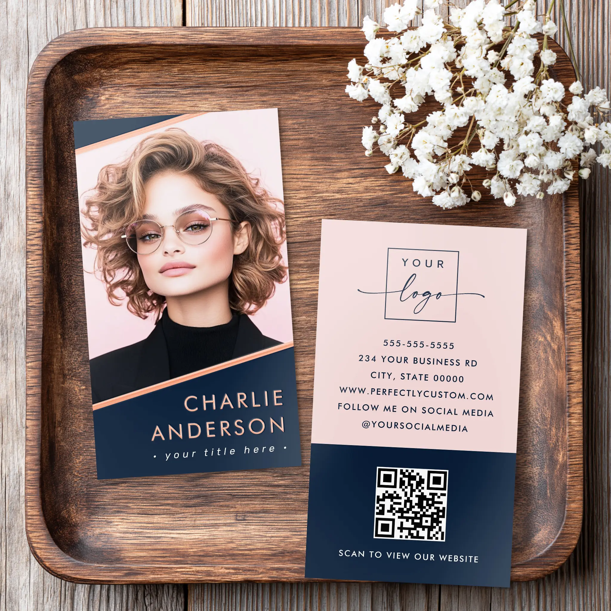Front and back view of a vertical business card. The front showcases a central custom portrait photo framed by diagonal navy blue color blocks and glowing rose gold foil stripe borders, with custom name and title below. The back features a blush pink top section with a custom logo and contact details, and a dark navy bottom section housing a scannable QR code.