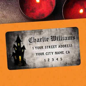 A rectangular self-adhesive return address label featuring a dark distressed charcoal parchment texture. On the left side is a dark silhouette illustration of a tall crooked haunted mansion with glowing yellow windows. On the right side is four lines of black text, featuring a top name line in gothic blackletter font followed by address lines in a vintage serif typeface.