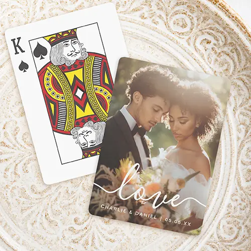 Playing card design featuring a full-bleed custom picture overlayed at the bottom with white hand-lettered calligraphic script reading love, and customizable uppercase sans-serif text below displaying names and a date separated by a vertical bar.
